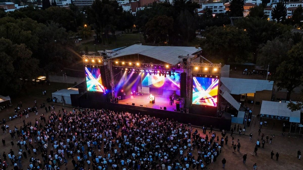 Imagen ilustrativa: vista aérea de un escenario de festival al aire libre en un parque al atardecer, con luces de colores y público lejano