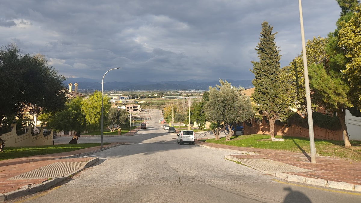 Residential street on a slope with streetlights, parked cars, and mountains in the background under dark clouds.