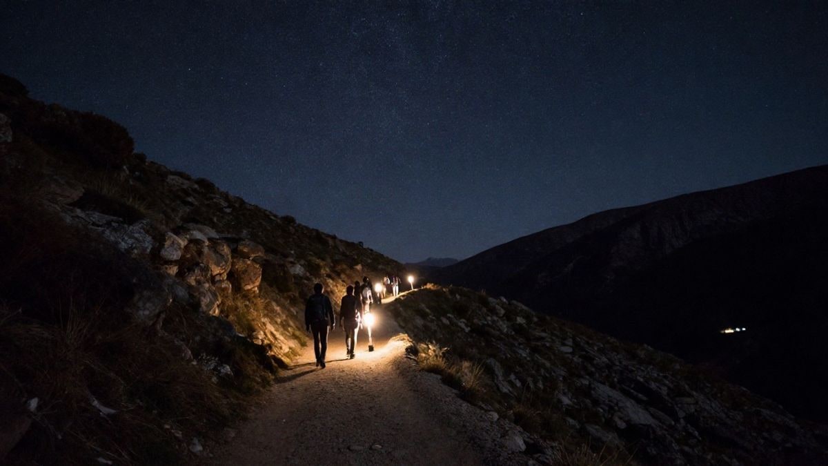 Illustrative image: night trail in the mountains with a group of hikers from behind under a starry sky