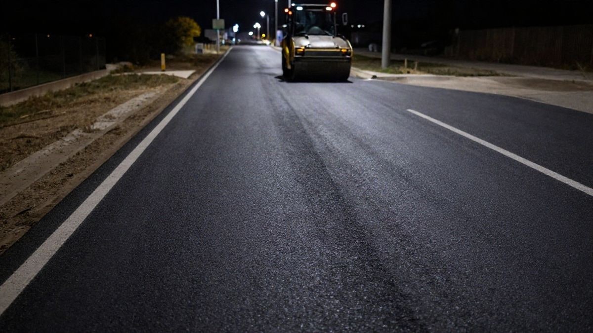 Imagen ilustrativa: carretera recién asfaltada de noche con luces de obra y una apisonadora al fondo