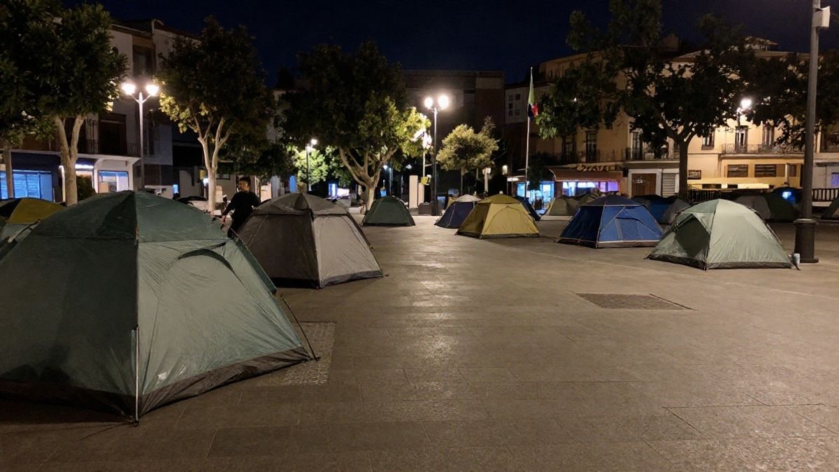 Imagen ilustrativa: tiendas de campaña dispersas en una plaza urbana iluminada por farolas durante la noche