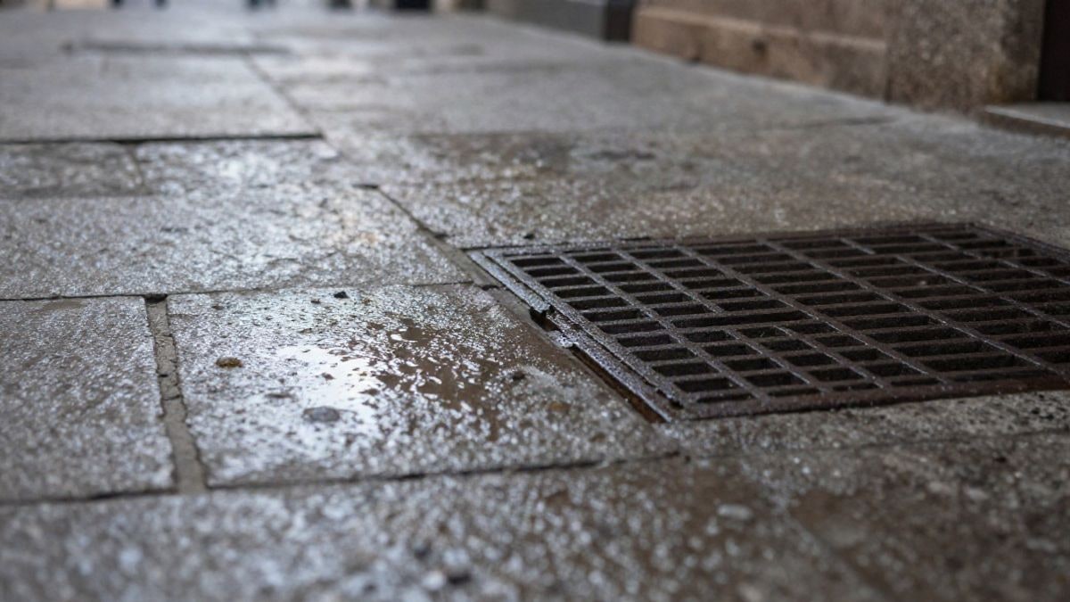 Illustrative image: wet stone pavement with accumulated water and a metal drainage grate at sunset