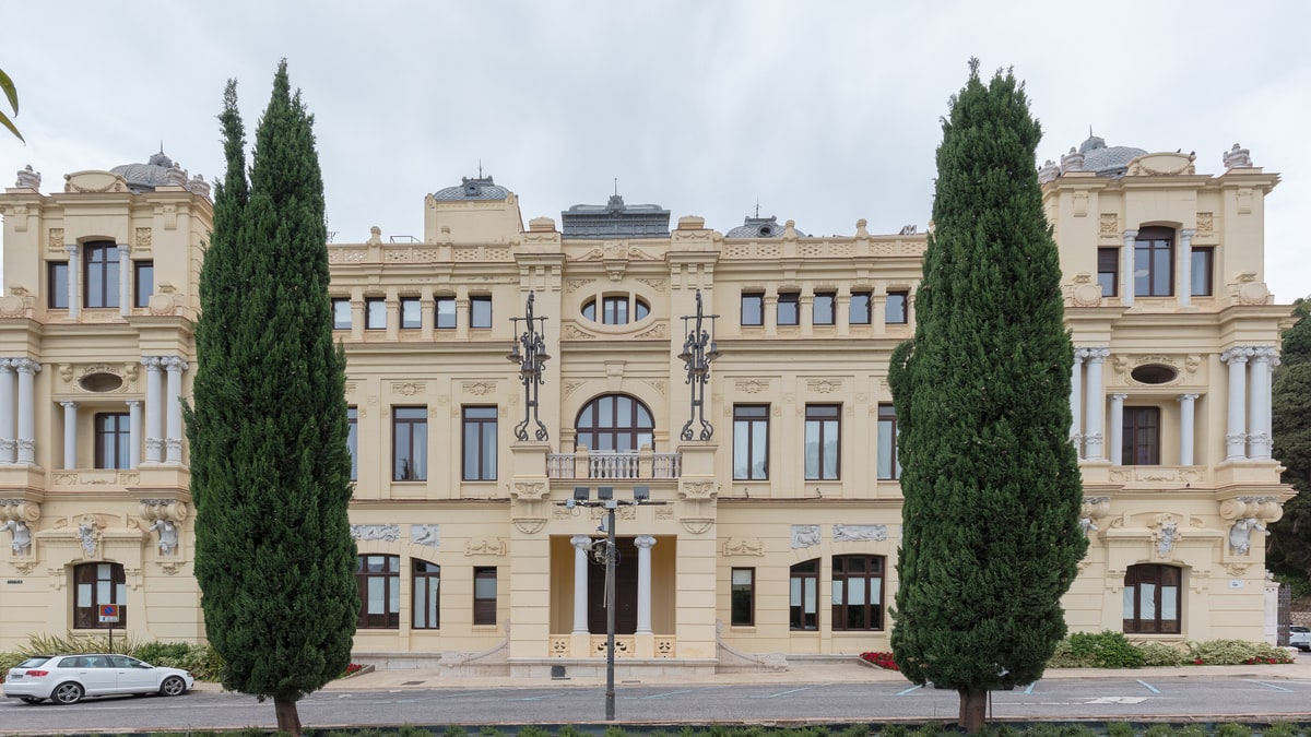 Fachada amarilla del Ayuntamiento de Málaga con cipreses y balcón central, vista frontal diurna.